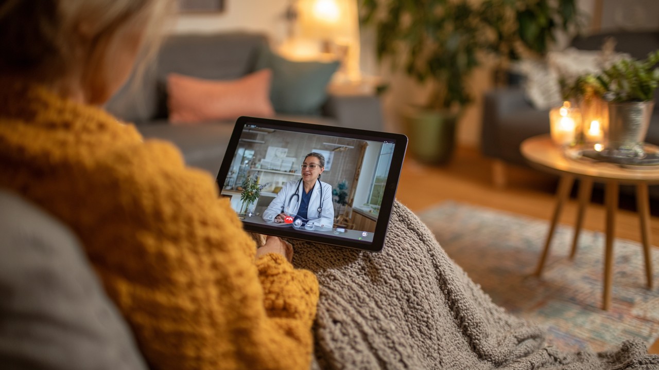 Patient consulting with doctor via video call on tablet at home, illustrating telemedicine and remote healthcare access in a cozy living room.