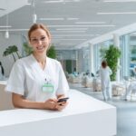 Modern sağlık kliniği resepsiyon alanında güler yüzlü resepsiyonist akıllı telefon kullanırken, doğal ışık ve sağlık posterleriyle aydınlık ve profesyonel ortam.