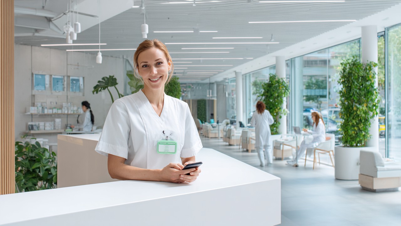 Modern sağlık kliniği resepsiyon alanında güler yüzlü resepsiyonist akıllı telefon kullanırken, doğal ışık ve sağlık posterleriyle aydınlık ve profesyonel ortam.