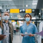 Modern havaalanında maske takmış çeşitli sağlık çalışanları, seyahat belgeleri ve COVID-19 güvenlik prosedürlerini tartışıyor.