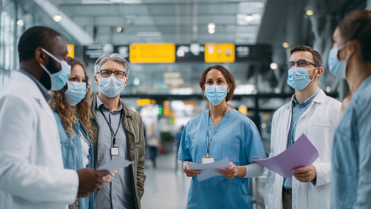 Modern havaalanında maske takmış çeşitli sağlık çalışanları, seyahat belgeleri ve COVID-19 güvenlik prosedürlerini tartışıyor.