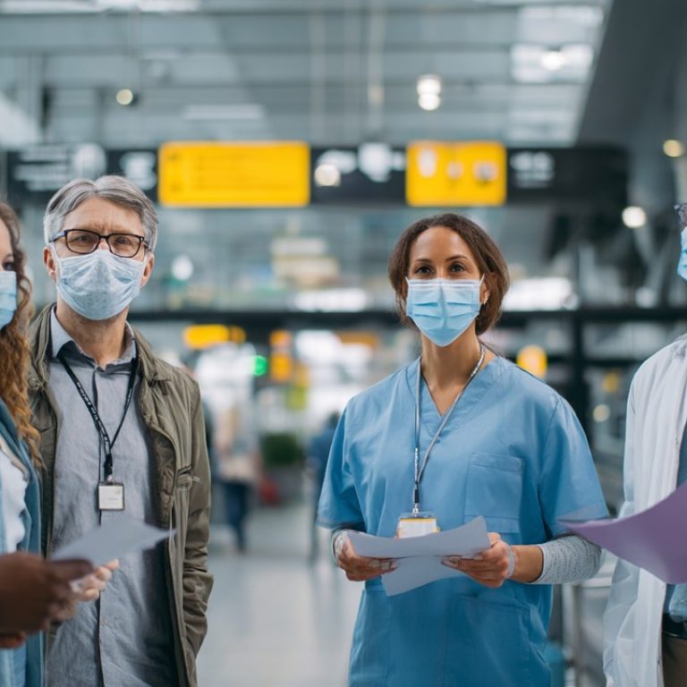 Modern havaalanında maske takmış çeşitli sağlık çalışanları, seyahat belgeleri ve COVID-19 güvenlik prosedürlerini tartışıyor.